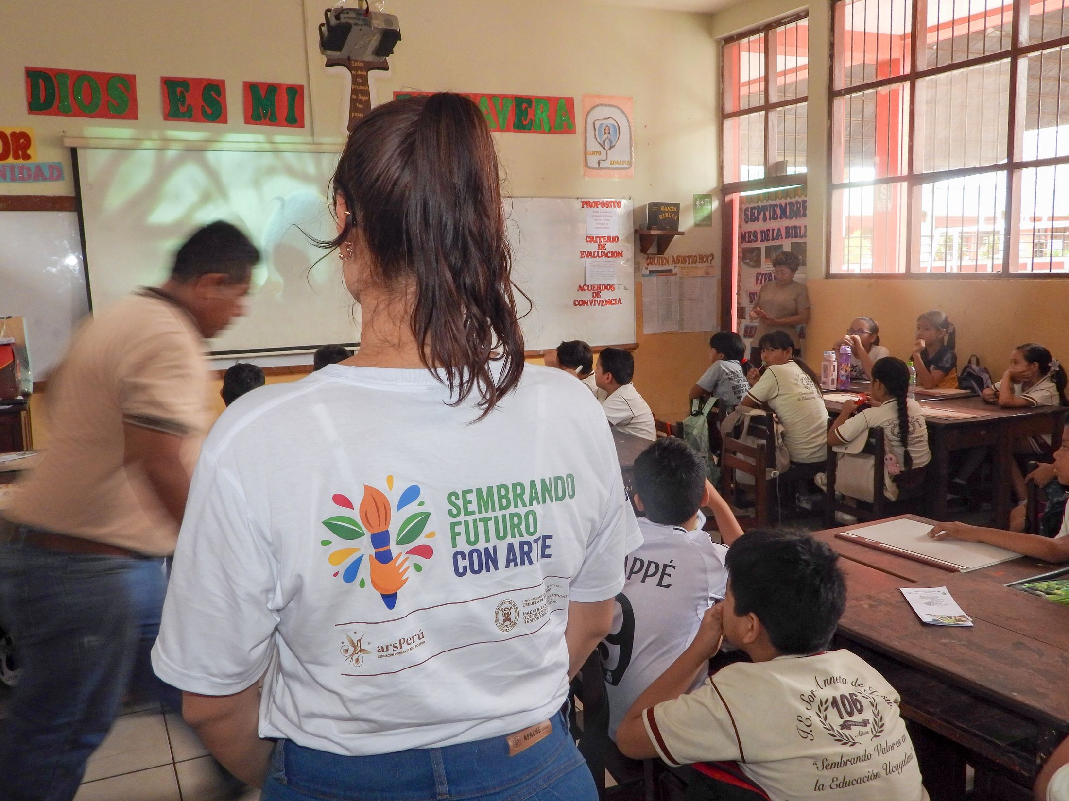 Integrante del equipo de ArsPerú, con la camiseta de la campaña Sembrando Futuro con Arte, frente a un aula de estudiantes en Pucallpa